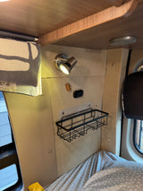 Inside an rv rental: a cozy sleeping nook with a wall-mounted reading lamp, a small shelf, and a patterned bed.