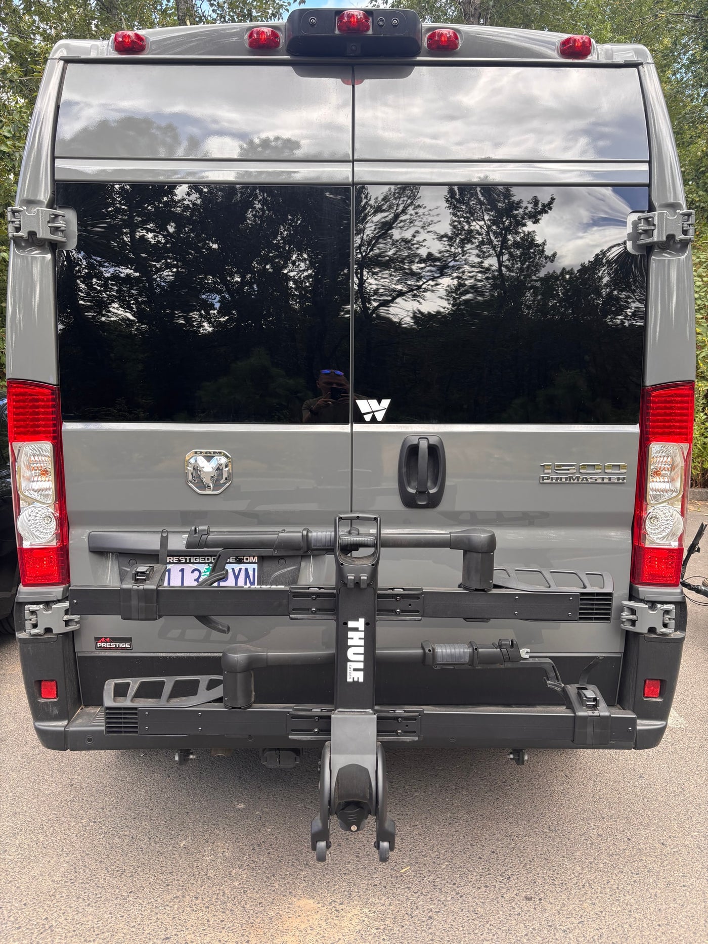 Rear view of a grey Ram ProMaster RV rental van equipped with a Thule bike rack. Dark windows reflect trees. — photo 9
