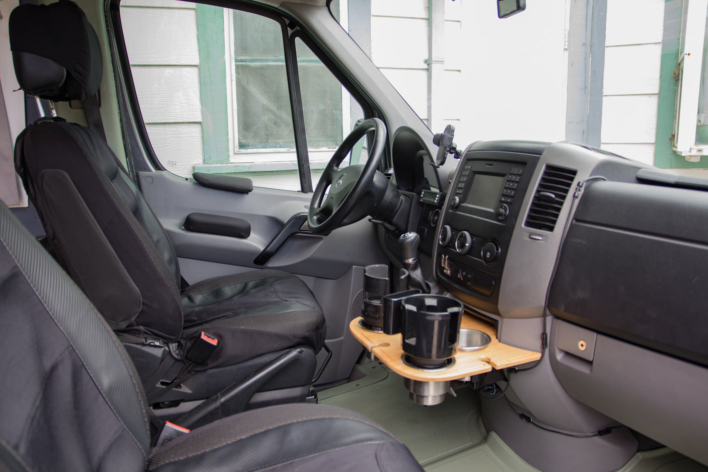 View of an RV rental's driver cockpit: seats, dashboard, custom wood cup holder tray, and infotainment.