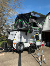 This RV rental, a teardrop-style camping trailer with a deployed black and green rooftop tent, is parked in a driveway.