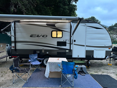 **RV rental** campsite with extended awning, lit interior window, table, chairs, and blue mat.