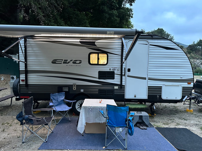 **RV rental** campsite with extended awning, lit interior window, table, chairs, and blue mat.