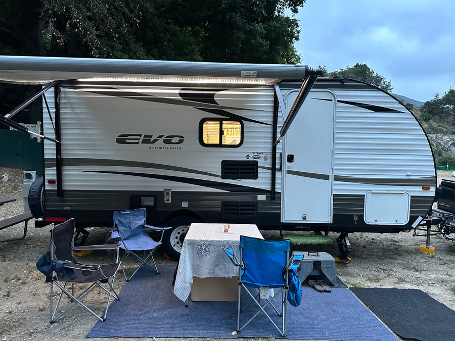 **RV rental** campsite with extended awning, lit interior window, table, chairs, and blue mat.