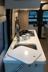 Inside an RV rental, a compact kitchen features a sink with a faucet, a two-burner stove, and two camping mugs.