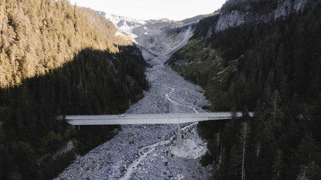 A white RV rental drives on a high bridge spanning a wide, rocky valley with a winding river and dense evergreen forests. Adventure awaits!
