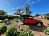 Red Jeep Wrangler with grey rooftop tent, ladder extended, set up at a sunny outdoor camp. Perfect for an RV rental trip.