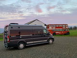 Dark gray RV rental parked on gravel near an old red double-decker bus and a white structure under a vibrant sky.