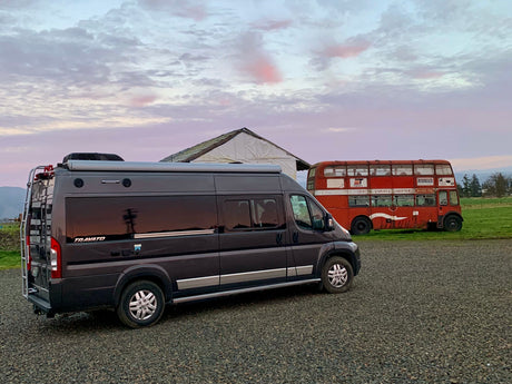 Dark gray RV rental parked on gravel near an old red double-decker bus and a white structure under a vibrant sky.