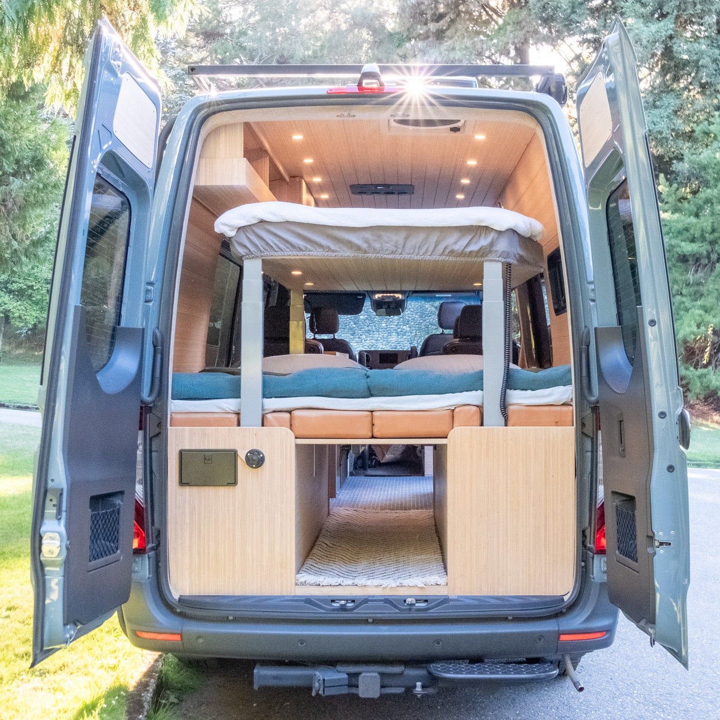 RV rental: Open rear doors of this custom camper van show its cozy interior, featuring elevated & lower beds, and wood paneling.
