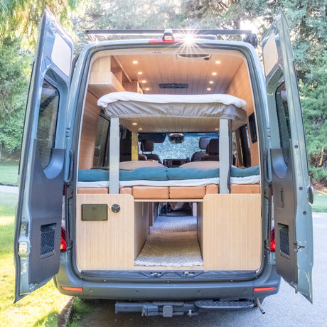 RV rental: Open rear doors of this custom camper van show its cozy interior, featuring elevated & lower beds, and wood paneling.