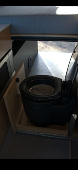 Inside this RV rental, a black pull-out composting toilet is shown on its sliding tray, with a bed overhead.