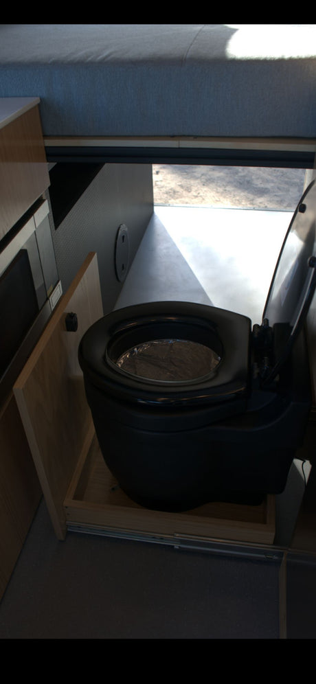 Inside this RV rental, a black pull-out composting toilet is shown on its sliding tray, with a bed overhead.