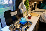 Compact RV rental kitchen with stove, kettle, sink, and comfy bed beyond. Perfect for mobile living.