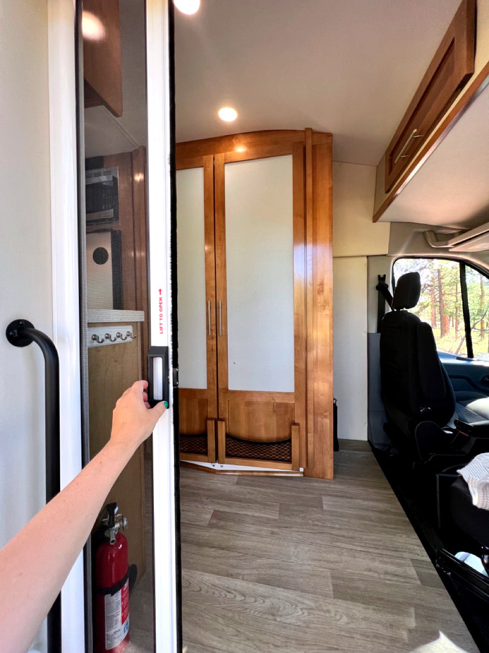 Interior of an RV rental showing a hand opening a screen door, cabinets, and a fire extinguisher, offering compact comfort.
