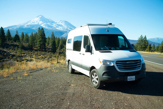 White RV rental van parked on a gravel shoulder with snow-capped mountains and pine trees under a clear blue sky.|A white RV rental van