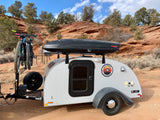 Teardrop trailer RV rental for desert adventures, with bikes and cargo box, ready for exploration in a red landscape.