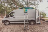 White RV rental van on sandy ground with surfboards, ladder, and a beach shower setup. Ready for coastal adventures!