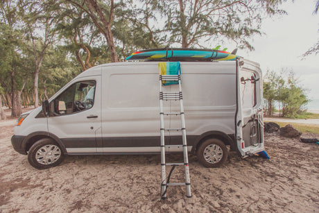 White RV rental van on sandy ground with surfboards, ladder, and a beach shower setup. Ready for coastal adventures!