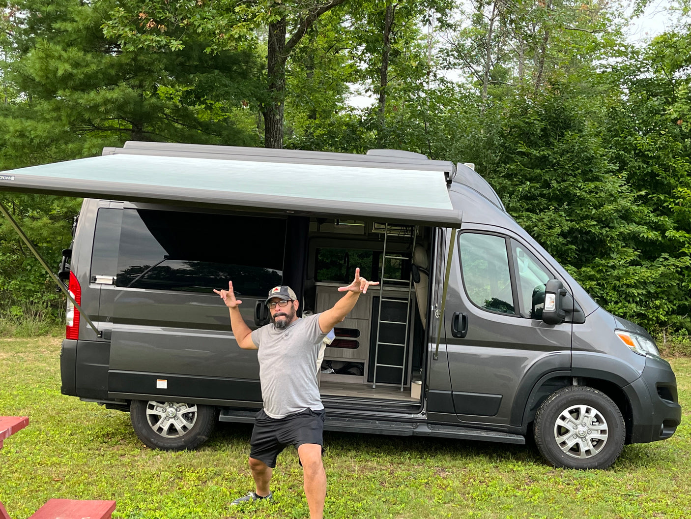 A man poses before a gray RV rental camper van with an extended awning. It's parked in a grassy area, ready for outdoor adventure.