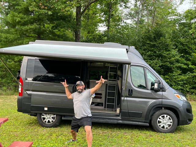 A man poses before a gray RV rental camper van with an extended awning. It's parked in a grassy area, ready for outdoor adventure.