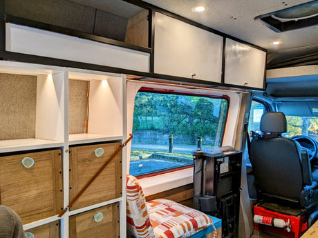 Bright RV rental interior: custom wooden storage, white overhead cabinets, a large window view, and a fire extinguisher.