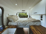 Cozy RV rental interior with a bed, white walls, wood counters, and a vivid green nature view from the back.
