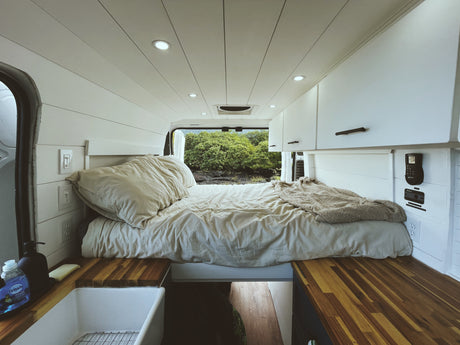 Cozy RV rental interior with a bed, white walls, wood counters, and a vivid green nature view from the back.