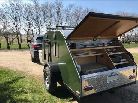 Green teardrop RV rental attached to a car, with its rear hatch open revealing a compact outdoor kitchen and storage.