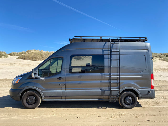 A dark gray camper van, perfect for an RV rental, parked on a sunny sandy beach with tall dunes under a clear blue sky.