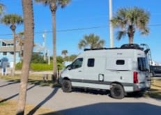  parked at a scenic lakeside campground.|Enjoy a stunning beach sunset from your rv rental! A camper van sits nestled among dunes & palms under a colorful sky.|Illuminated rv rental camper van with awning & underglow lights at night — photo 7
