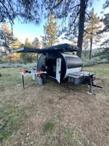 This off-grid teardrop camper, perfect for RV rental adventures, features an outdoor kitchen and solar panels in a forest setting.
