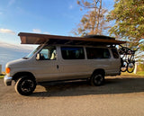 Tan adventure campervan with extended wooden roof, cargo box, and bikes parked by the ocean. Perfect for an RV rental trip.