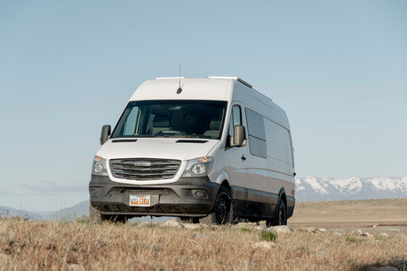 A white Freightliner camper van, likely for RV rental, is on a dry, grassy Utah hill with snow-capped mountains.