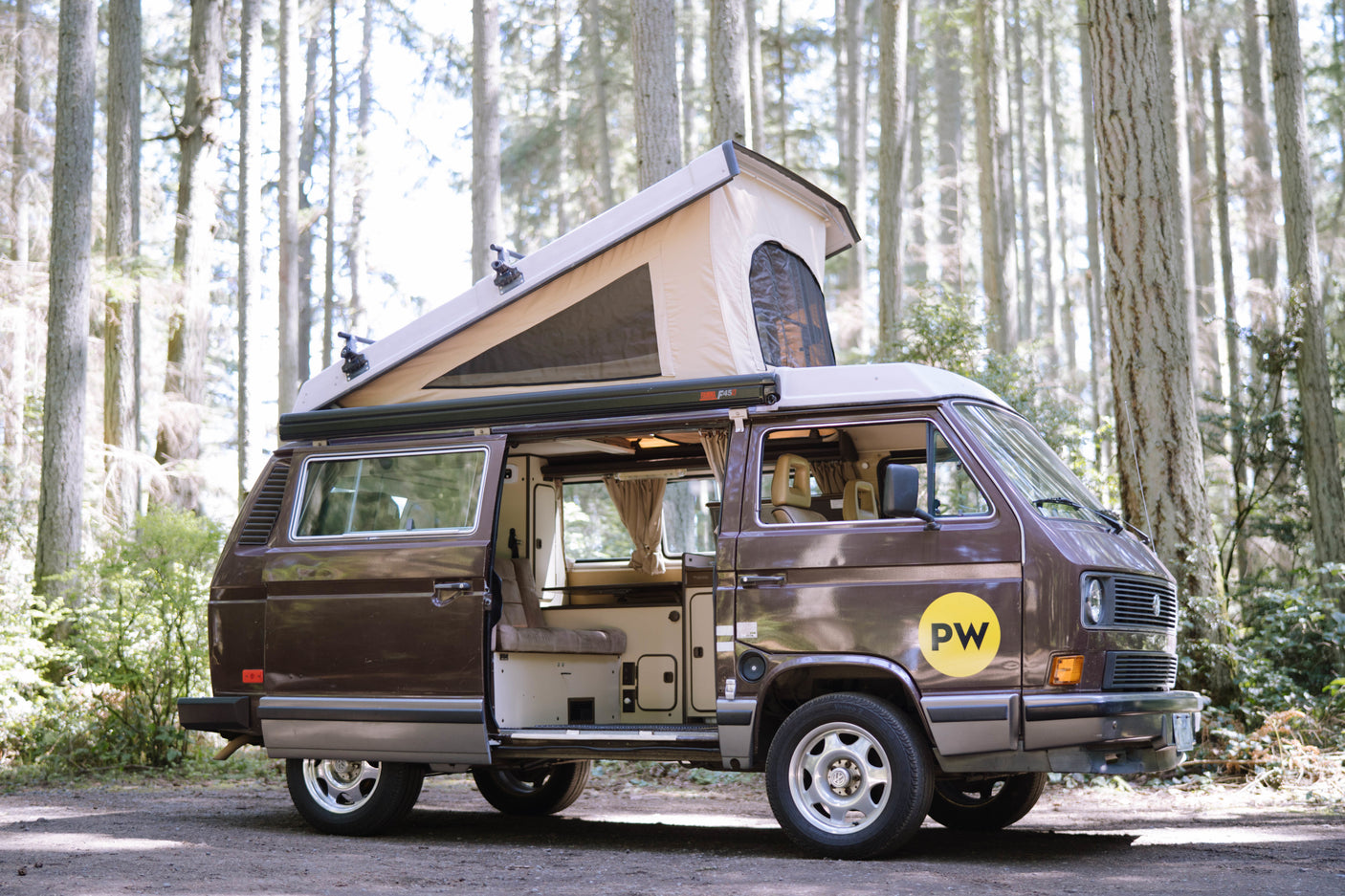 A brown VW pop-top camper van, with its door open revealing the cozy interior, parked in a sunny forest. Perfect for RV rental.