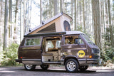 A brown VW pop-top camper van, with its door open revealing the cozy interior, parked in a sunny forest. Perfect for RV rental.