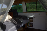 This rv rental shows a cozy bed, cabinet storage with snacks, and windows overlooking green nature.