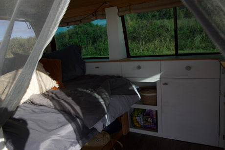 This rv rental shows a cozy bed, cabinet storage with snacks, and windows overlooking green nature.