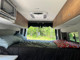 Cozy RV rental interior: large bed with dark comforter, overhead microwave/AC, and lush forest view from the back.