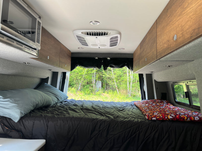 Cozy RV rental interior: large bed with dark comforter, overhead microwave/AC, and lush forest view from the back. — photo 14