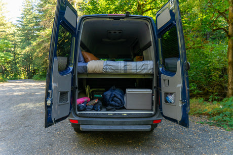 Open rear doors of a blue RV rental reveal a comfortable bed above and ample gear storage below for your next van life adventure.