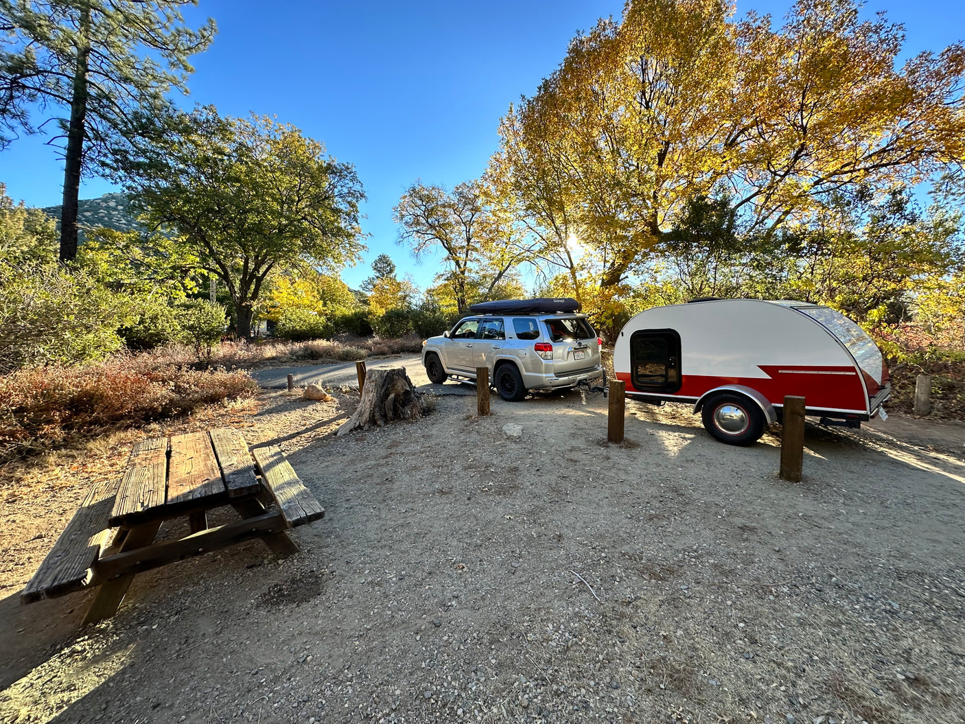Autumn RV rental: Teardrop trailer & SUV at a sunlit campsite with a picnic table.