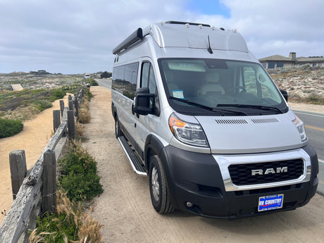 Sleek silver camper van RV rental parked on a scenic coastal trail alongside a rustic fence. Cloudy skies over distant homes.