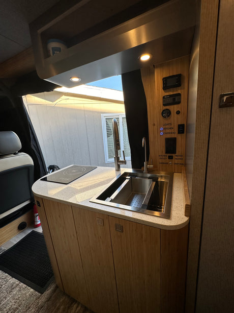Modern RV rental kitchen featuring wood cabinetry, white countertop, sink, induction cooktop, and a detailed control panel.