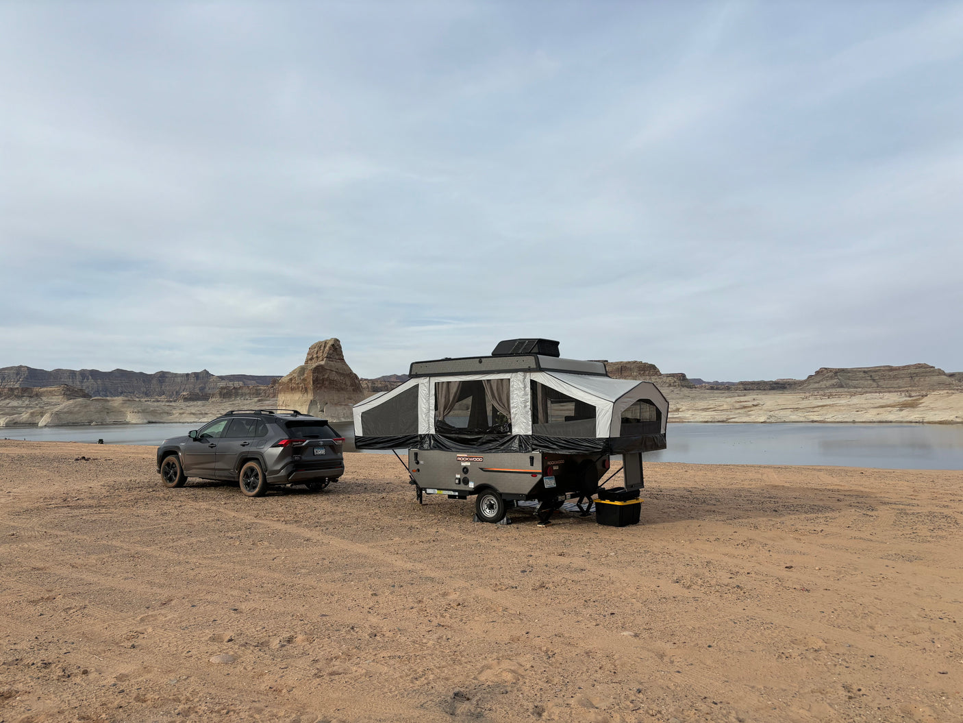 An RV rental pop-up camper and SUV are parked on sandy terrain beside a tranquil lake with layered rock cliffs.