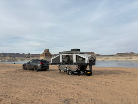 An RV rental pop-up camper and SUV are parked on sandy terrain beside a tranquil lake with layered rock cliffs.