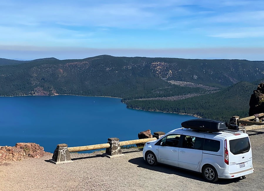 White RV rental van parked at a stunning lake viewpoint with dense forests and mountains under a blue sky.