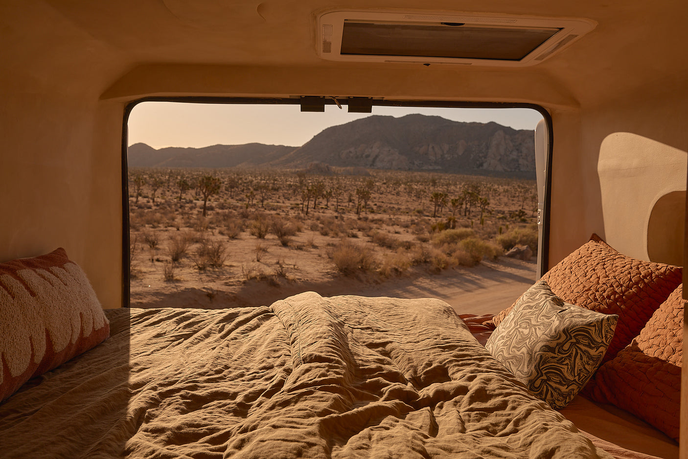 Inside an rv rental: a comfy bed with warm blankets, framing a window view of a desert, Joshua trees, & mountains.