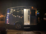 Silver RV rental illuminated by string lights at night, with its door open. Experience a cozy camping adventure.