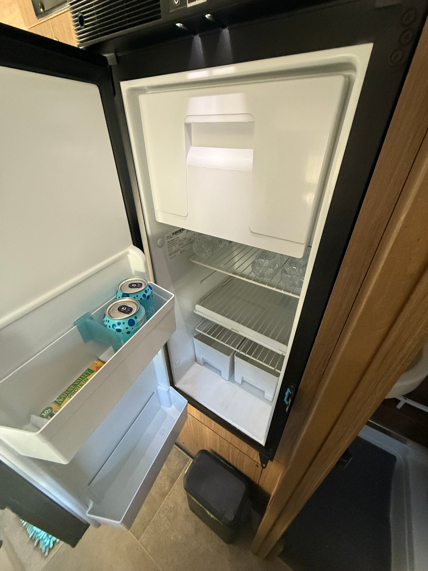 An open compact refrigerator inside an RV rental, revealing interior shelves with water bottles, cans, and a protein bar.