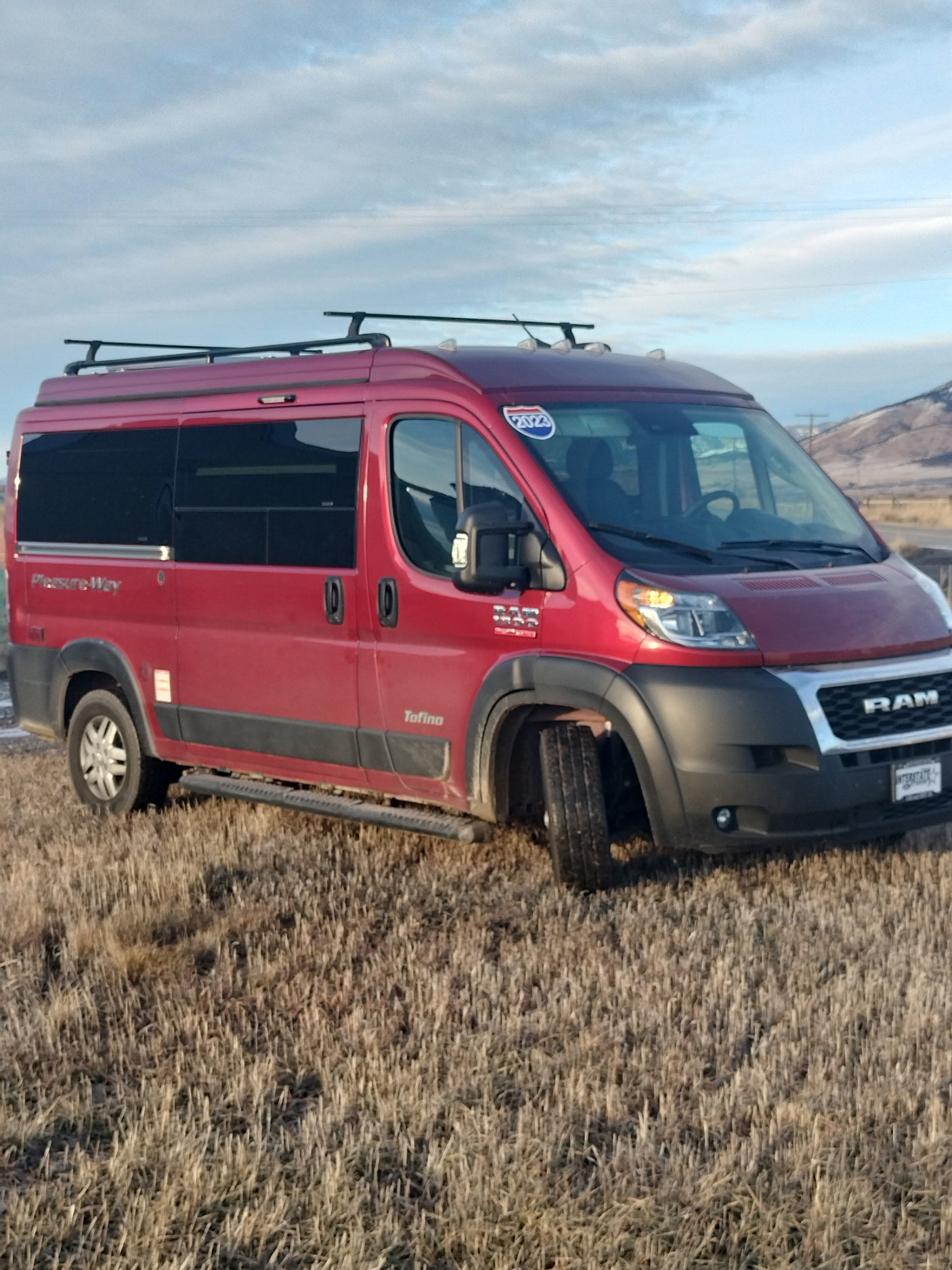 Red Pleasure Way rv rental van (Ram ProMaster) with roof rack, parked in a dry field under a blue sky.